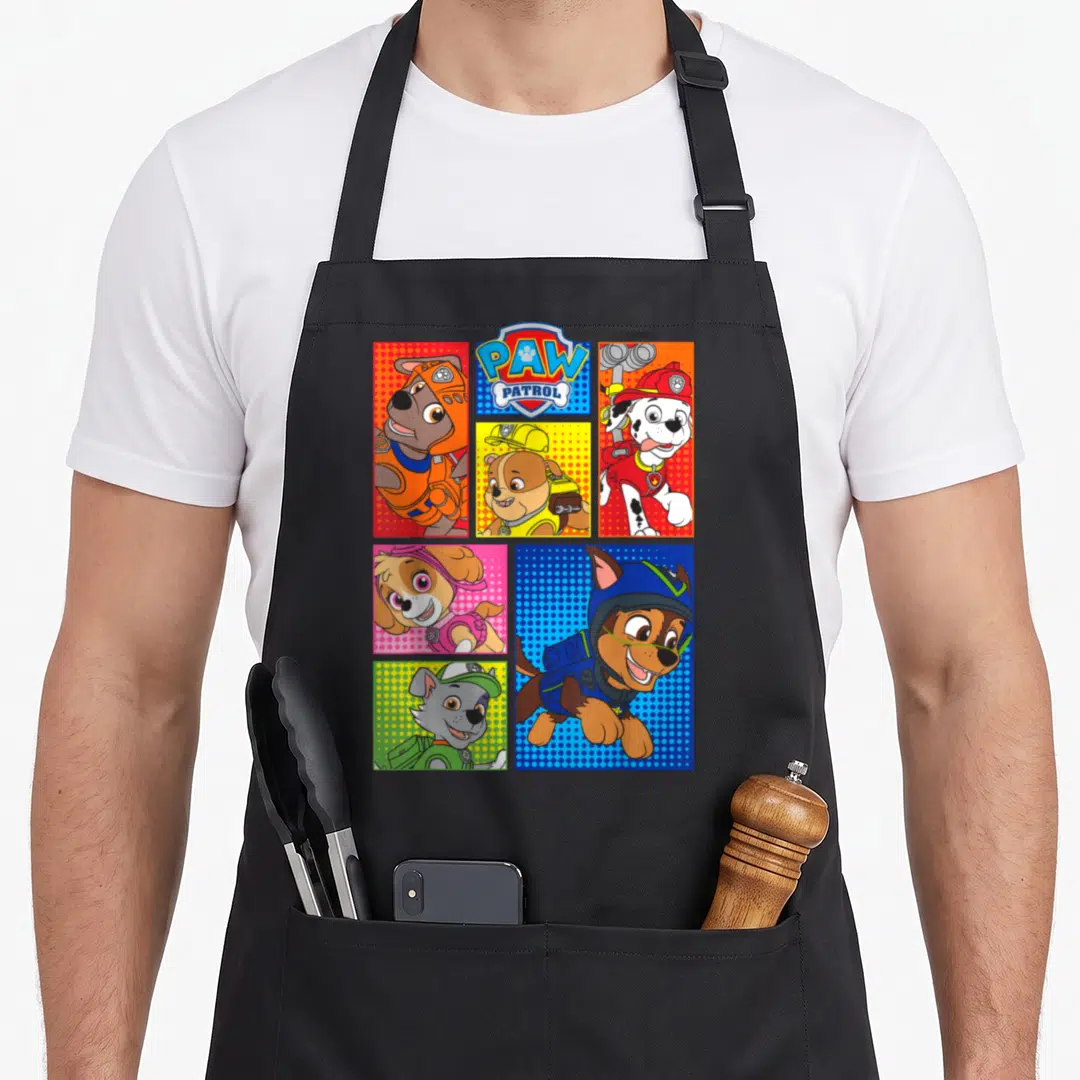 Pup novelty apron - Five panel grid of cartoon puppies with PAW PATROL logo in blue red yellow and orange — shown on a black cotton canvas apron against a white background