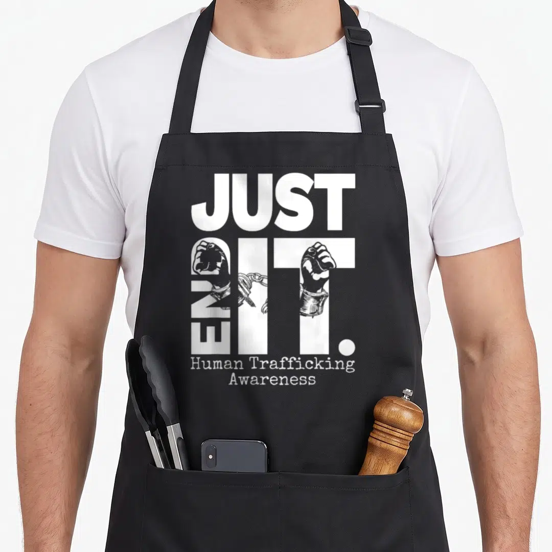Funny end human trafficking awareness apron - White JUST END IT typography with shackled hands filling the letters — shown on a black cotton canvas apron against a white background