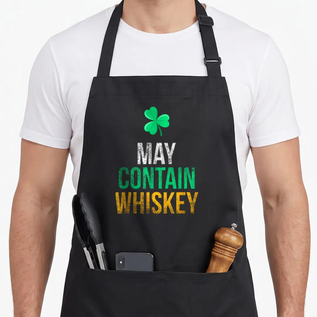 May funny irish aprons - Green shamrock above distressed MAY, CONTAIN, WHISKEY text in white, green, and orange — shown on a black cotton canvas apron against a white background