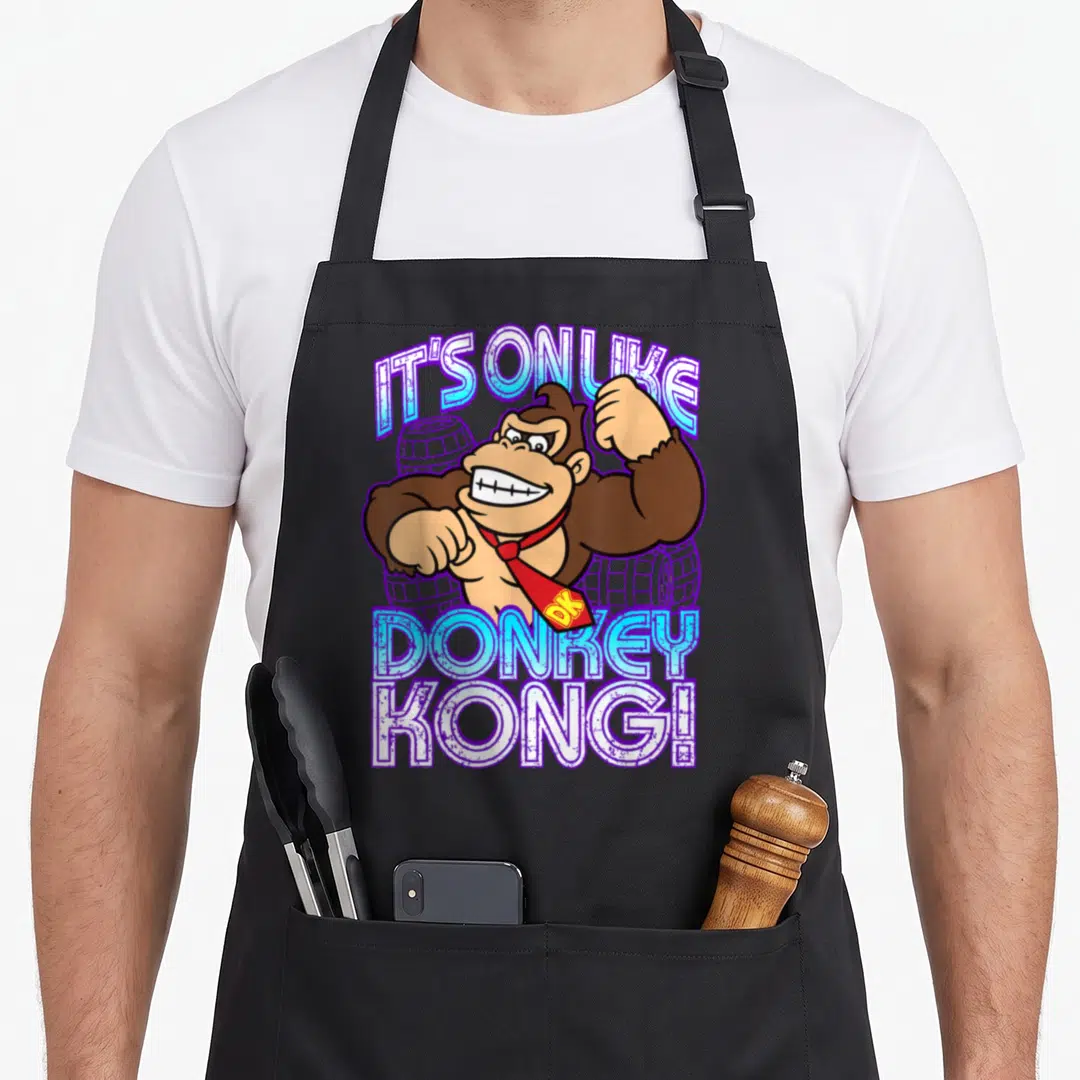 Taunt donkey apron - Brown cartoon gorilla flexing with It's On Like Donkey Kong text and purple barrels — shown on a black cotton canvas apron against a white background