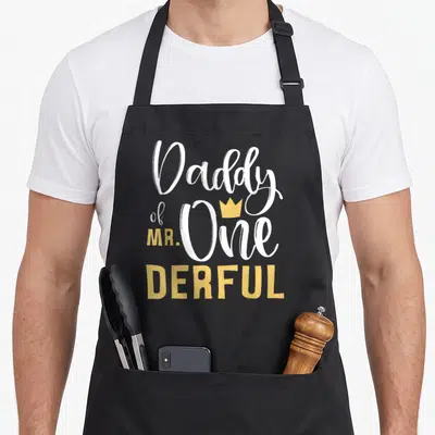 Onederful daddy apron - Daddy of MR One Derful text with small yellow crown above the word One — shown on a black cotton canvas apron against a white background