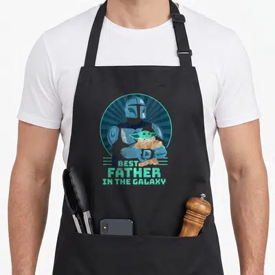 Funny father day apron - Mandalorian holding Grogu in blue-grey sunburst with mint green text best father in the galaxy — shown on a black cotton canvas apron against a white background