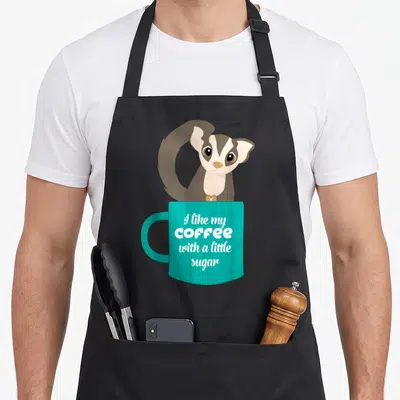 Sugar coffee apron - Cartoon sugar glider peeking from behind teal mug with white stacked text — shown on a black cotton canvas apron against a white background