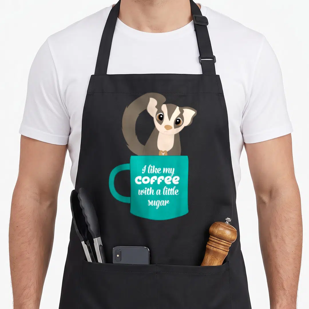 Sugar coffee apron - Cartoon sugar glider peeking from behind teal mug with white stacked text — shown on a black cotton canvas apron against a white background