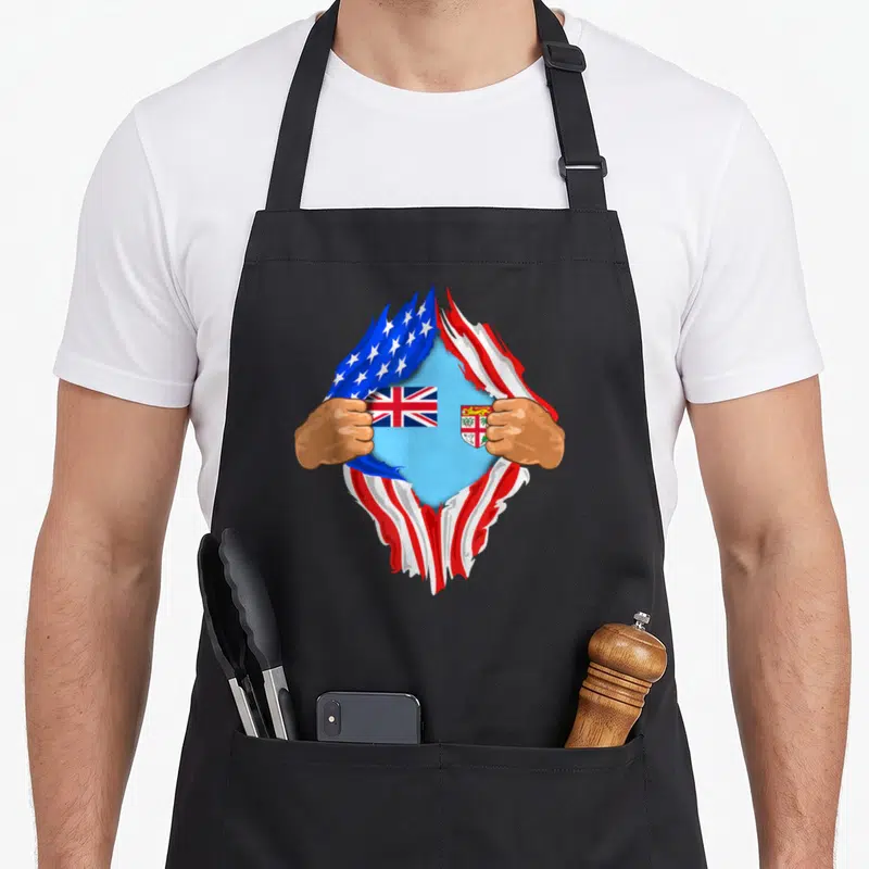 Fijian blood apron - Two brown hands tearing USA flag to reveal light blue Fiji flag with shield emblem — shown on a black cotton canvas apron against a white background