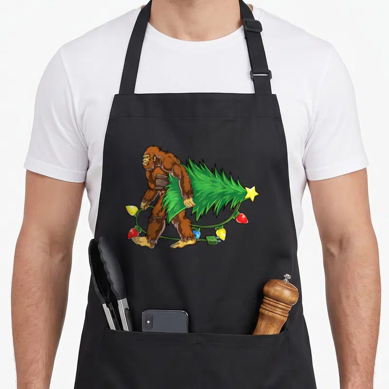 Bigfoot christmas tree apron - Cartoon brown Bigfoot walking carrying green Christmas tree and colorful lights — shown on a black cotton canvas apron against a white background