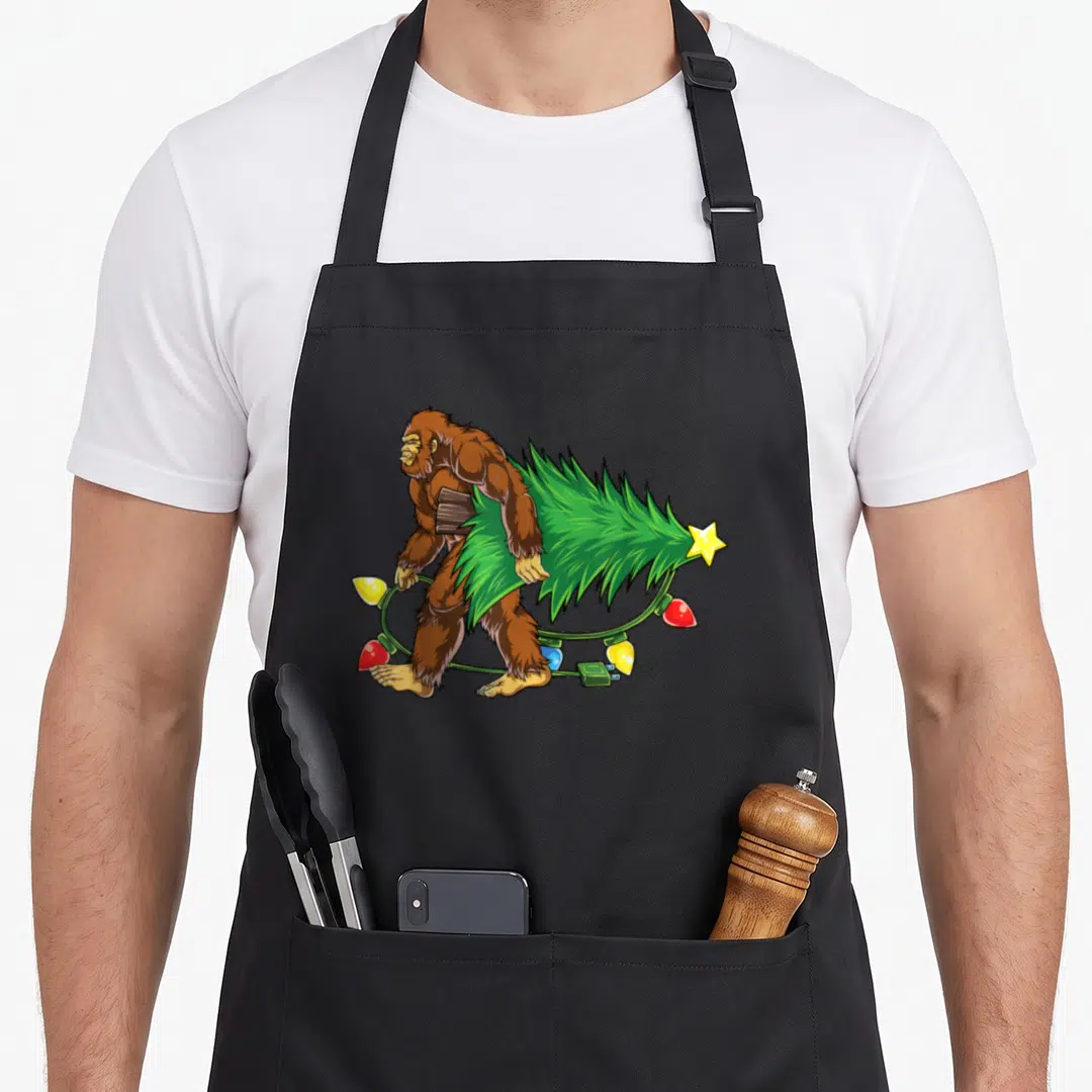 Bigfoot christmas tree apron - Cartoon brown Bigfoot walking carrying green Christmas tree and colorful lights — shown on a black cotton canvas apron against a white background