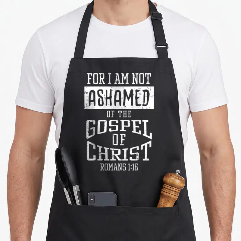Ashamed bible verse apron - White distressed typography reading For I Am Not Ashamed Of The Gospel Of Christ Romans — shown on a black cotton canvas apron against a white background
