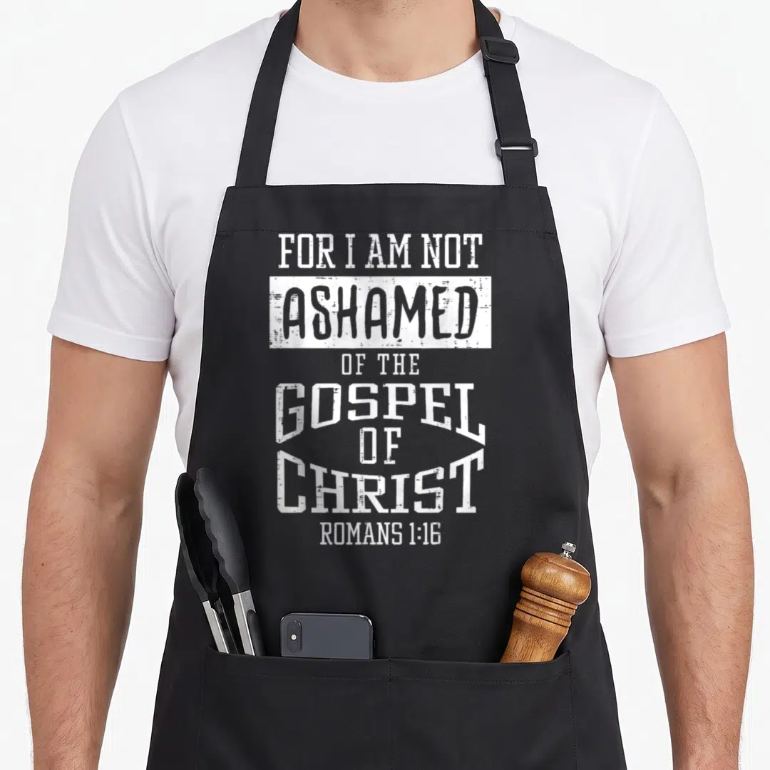 Ashamed bible verse apron - White distressed typography reading For I Am Not Ashamed Of The Gospel Of Christ Romans — shown on a black cotton canvas apron against a white background
