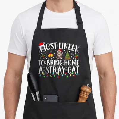 Most christmas cat apron - Cartoon cat in Santa hat with text Most Likely To Bring Home A Stray Cat surrounded by bells — shown on a black cotton canvas apron against a white background