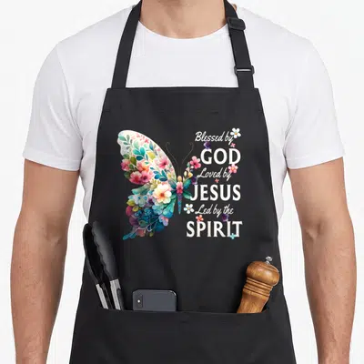 Loved floral apron - Watercolor butterfly with white, pink, blue, purple, and yellow flowers next to stacked religious — shown on a black cotton canvas apron against a white background