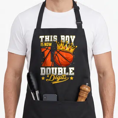 Ten double apron - Cartoon basketball forming number 10 with gold crown and bold text THIS BOY IS NOW 10 DOUBLE Digits — shown on a black cotton canvas apron against a white background