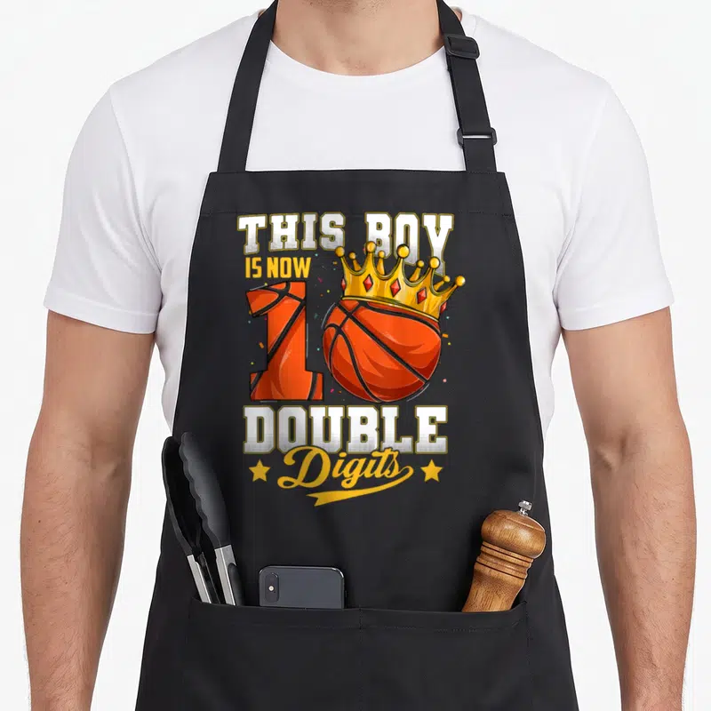 Ten double apron - Cartoon basketball forming number 10 with gold crown and bold text THIS BOY IS NOW 10 DOUBLE Digits — shown on a black cotton canvas apron against a white background