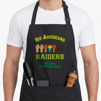 Rio americana apron - Seven raised fists including one rainbow fist below arched Rio Americano Raiders text — shown on a black cotton canvas apron against a white background