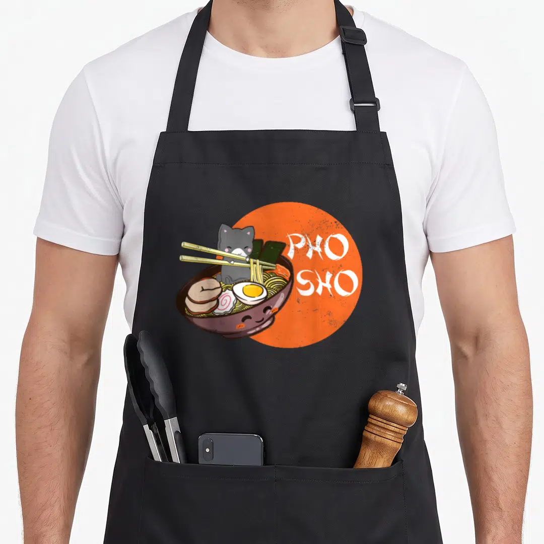 Pho cat apron - Grey cartoon cat holding chopsticks in noodle bowl with PHO SHO text on orange circle — shown on a black cotton canvas apron against a white background