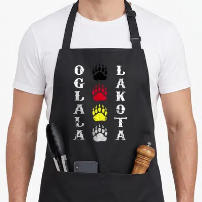 Oglala south boy apron - Four stacked bear paw prints in black, red, yellow, white with vertical OGLALA LAKOTA text — shown on a black cotton canvas apron against a white background