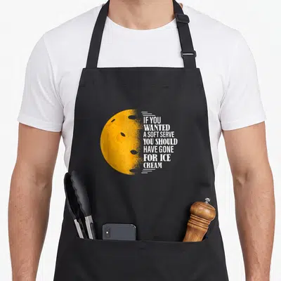 Wanted pickleball to serve man apron - Yellow moon with craters and white text reading If You Wanted A Soft Serve You — shown on a black cotton canvas apron against a white background