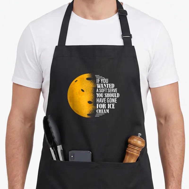 Wanted pickleball to serve man apron - Yellow moon with craters and white text reading If You Wanted A Soft Serve You — shown on a black cotton canvas apron against a white background