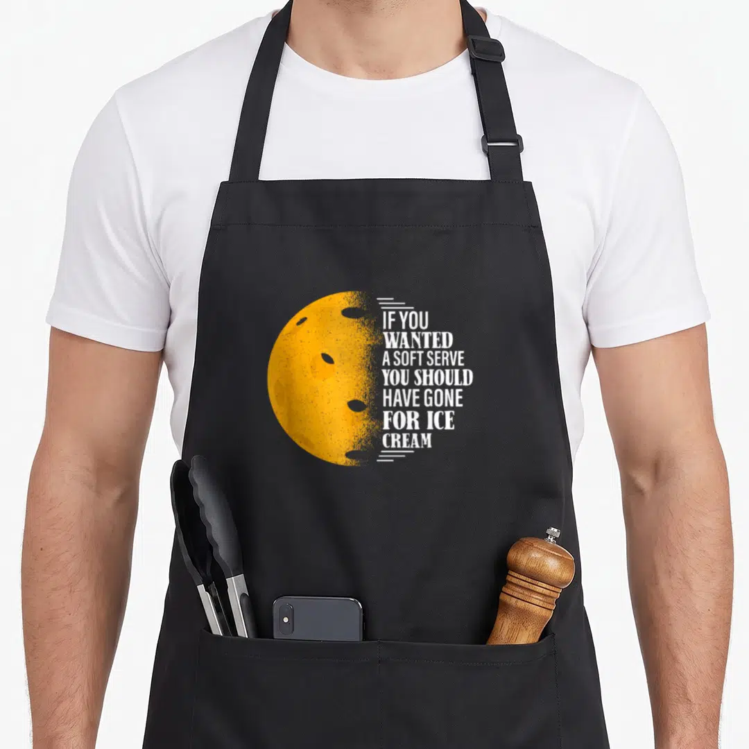 Wanted pickleball to serve man apron - Yellow moon with craters and white text reading If You Wanted A Soft Serve You — shown on a black cotton canvas apron against a white background