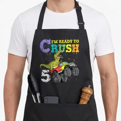5th dinosaur apron - Green T-Rex driving red and yellow monster truck with confetti and I'm Ready to Crush 5 text — shown on a black cotton canvas apron against a white background