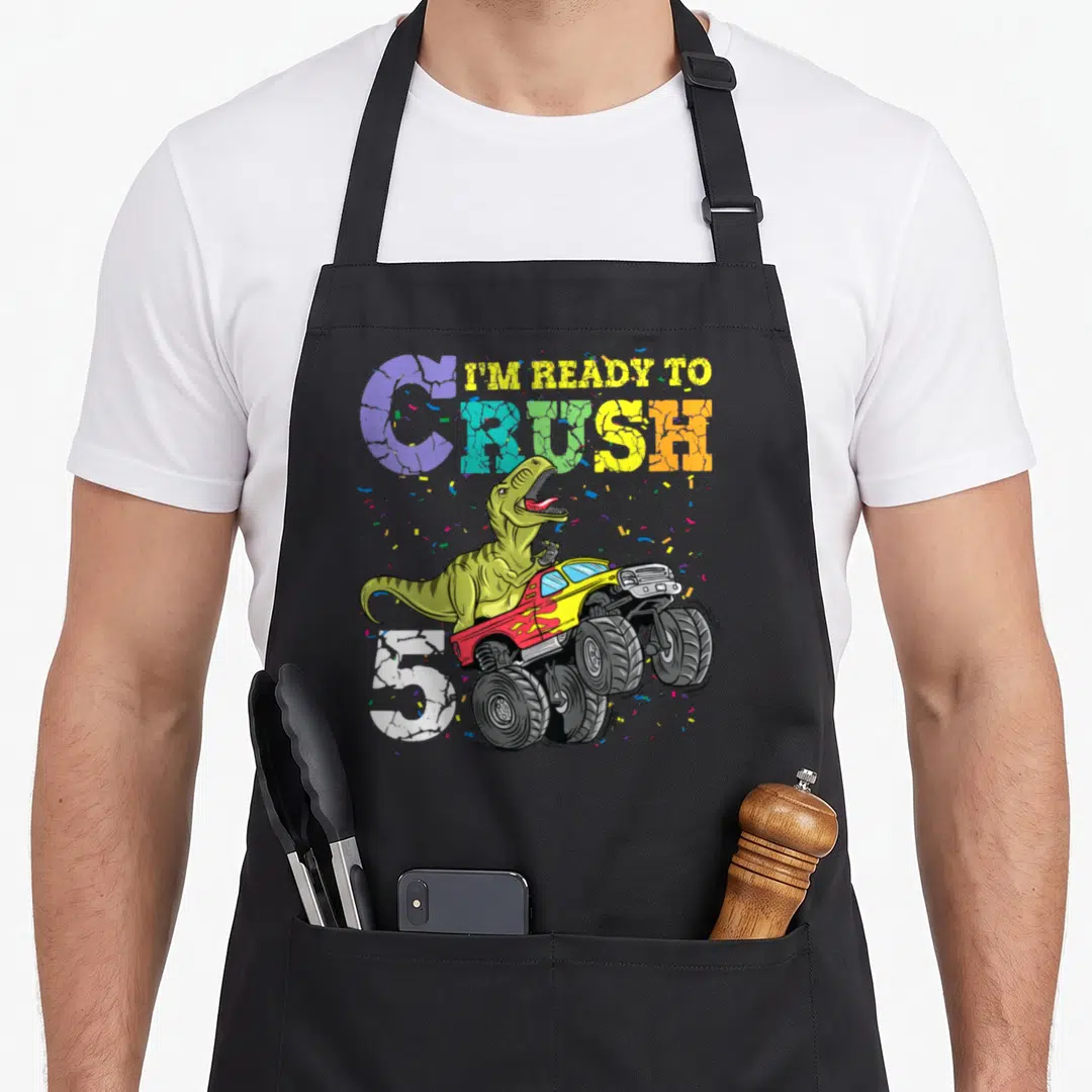 5th dinosaur apron - Green T-Rex driving red and yellow monster truck with confetti and I'm Ready to Crush 5 text — shown on a black cotton canvas apron against a white background