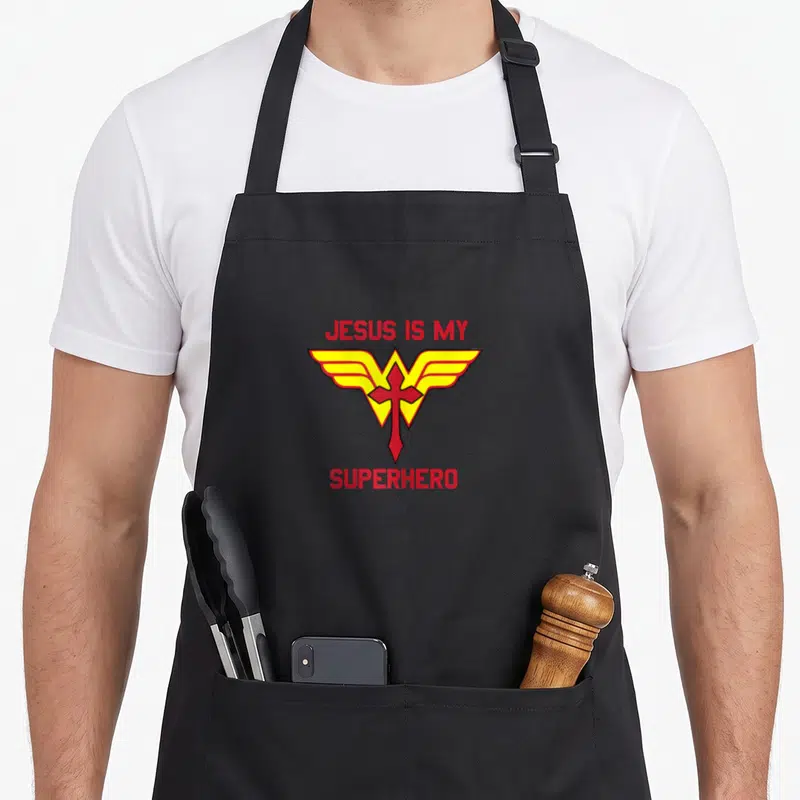 School christian apron - Red and yellow winged cross with JESUS IS MY SUPERHERO text — shown on a black cotton canvas apron against a white background