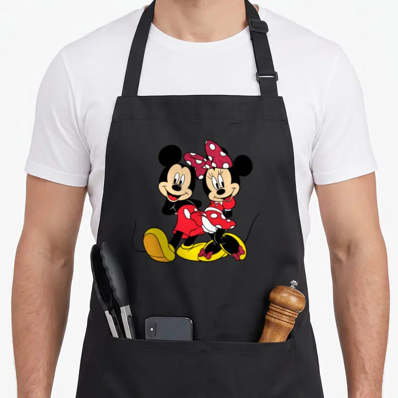 Funny mouse apron - Mickey and Minnie Mouse standing close together in a cartoon style wearing red and yellow outfits — shown on a black cotton canvas apron against a white background