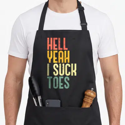 Hell inappropriate apron - Stacked text reading Hell Yeah I Suck Toes in red yellow beige and green distressed font — shown on a black cotton canvas apron against a white background