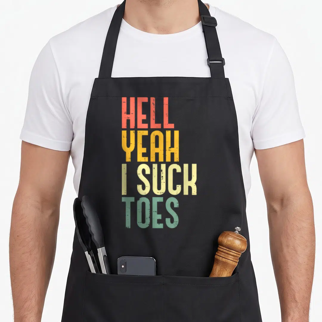 Hell inappropriate apron - Stacked text reading Hell Yeah I Suck Toes in red yellow beige and green distressed font — shown on a black cotton canvas apron against a white background