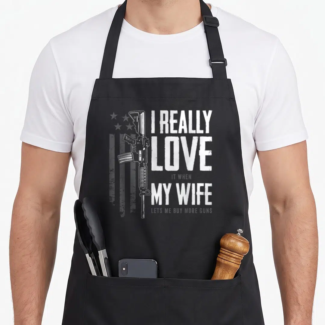 Funny gun wife apron - White distressed text I Really Love It When My Wife Lets Me Buy More Guns beside a rifle — shown on a black cotton canvas apron against a white background