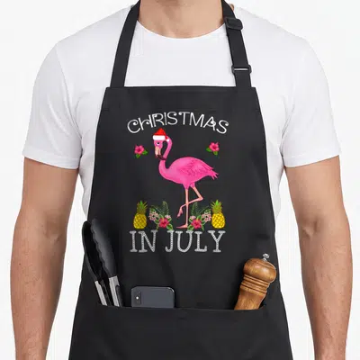 July pink flamingo apron - Cartoon pink flamingo in Santa hat with two pineapples and 'Christmas In July' text — shown on a black cotton canvas apron against a white background
