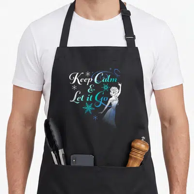 Nature novelty apron - Stacked Keep Calm & Let it Go text with standing Elsa and blue white snowflakes — shown on a black cotton canvas apron against a white background