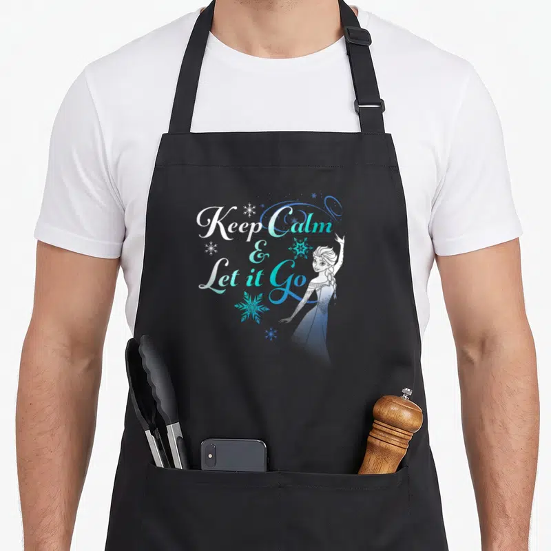 Nature novelty apron - Stacked Keep Calm & Let it Go text with standing Elsa and blue white snowflakes — shown on a black cotton canvas apron against a white background