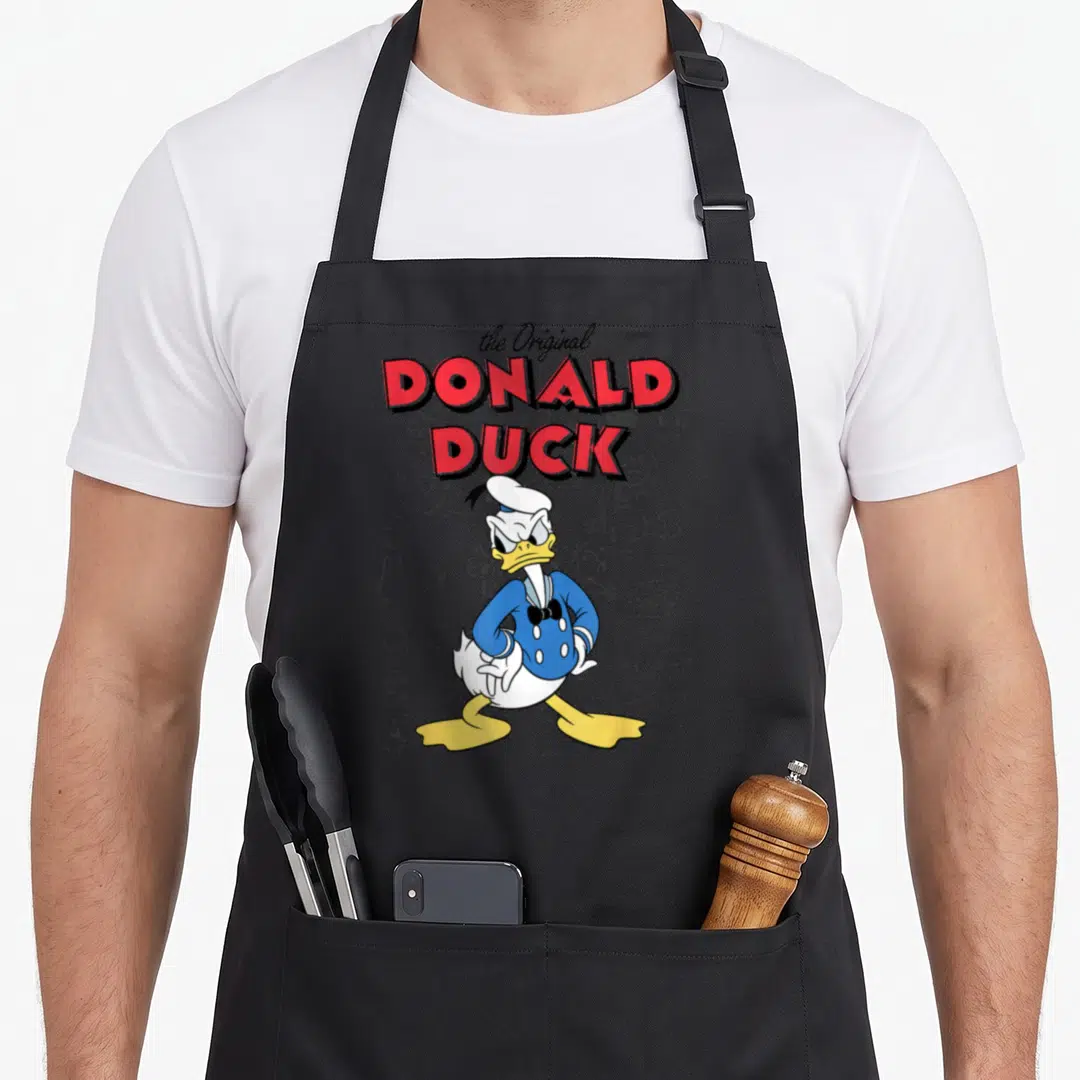 Sketch duck apron - Donald Duck standing over grey technical sketches with red and black stacked text — shown on a black cotton canvas apron against a white background