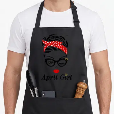 April custom face apron - Cartoon woman with red polka dot headband, glasses, and 'April Girl' text — shown on a black cotton canvas apron against a white background