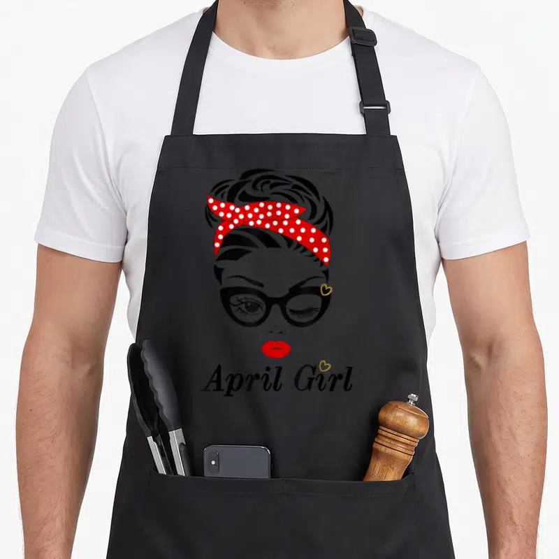 April custom face apron - Cartoon woman with red polka dot headband, glasses, and 'April Girl' text — shown on a black cotton canvas apron against a white background
