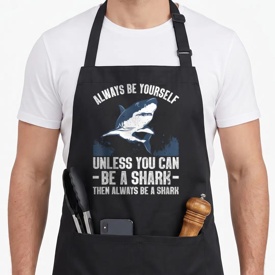 Biology megalodon apron - Distressed white and blue shark swimming over seaweed with text always be yourself unless you — shown on a black cotton canvas apron against a white background