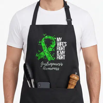 Gastroparesis wife apron - Green scattered hearts forming a ribbon next to white block text and script lettering — shown on a black cotton canvas apron against a white background