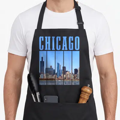 Illinois pride chicago apron - Blue CHICAGO text above a vertical strip photo of the city skyline in blue and grey tones — shown on a black cotton canvas apron against a white background