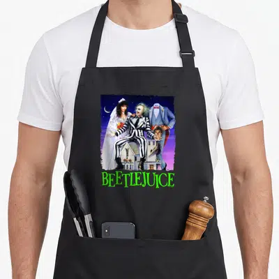 Title spooky apron - Beetlejuice collage with moon, house, and lime green text on purple background — shown on a black cotton canvas apron against a white background