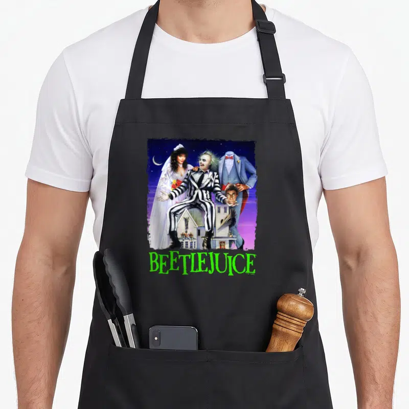 Title spooky apron - Beetlejuice collage with moon, house, and lime green text on purple background — shown on a black cotton canvas apron against a white background