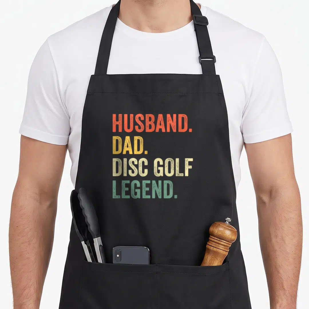 Disc father day apron - Stacked text HUSBAND. DAD. DISC GOLF. LEGEND. in orange, yellow, beige, and green — shown on a black cotton canvas apron against a white background