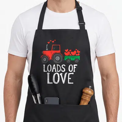 Load valentine's day apron - Red tractor pulling green trailer filled with hearts above stacked white text reading — shown on a black cotton canvas apron against a white background