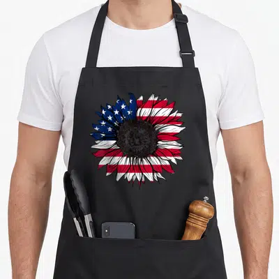 Flag sunflower 4th of july apron - Realistic sunflower with petals patterned in US flag blue white and red stripes — shown on a black cotton canvas apron against a white background