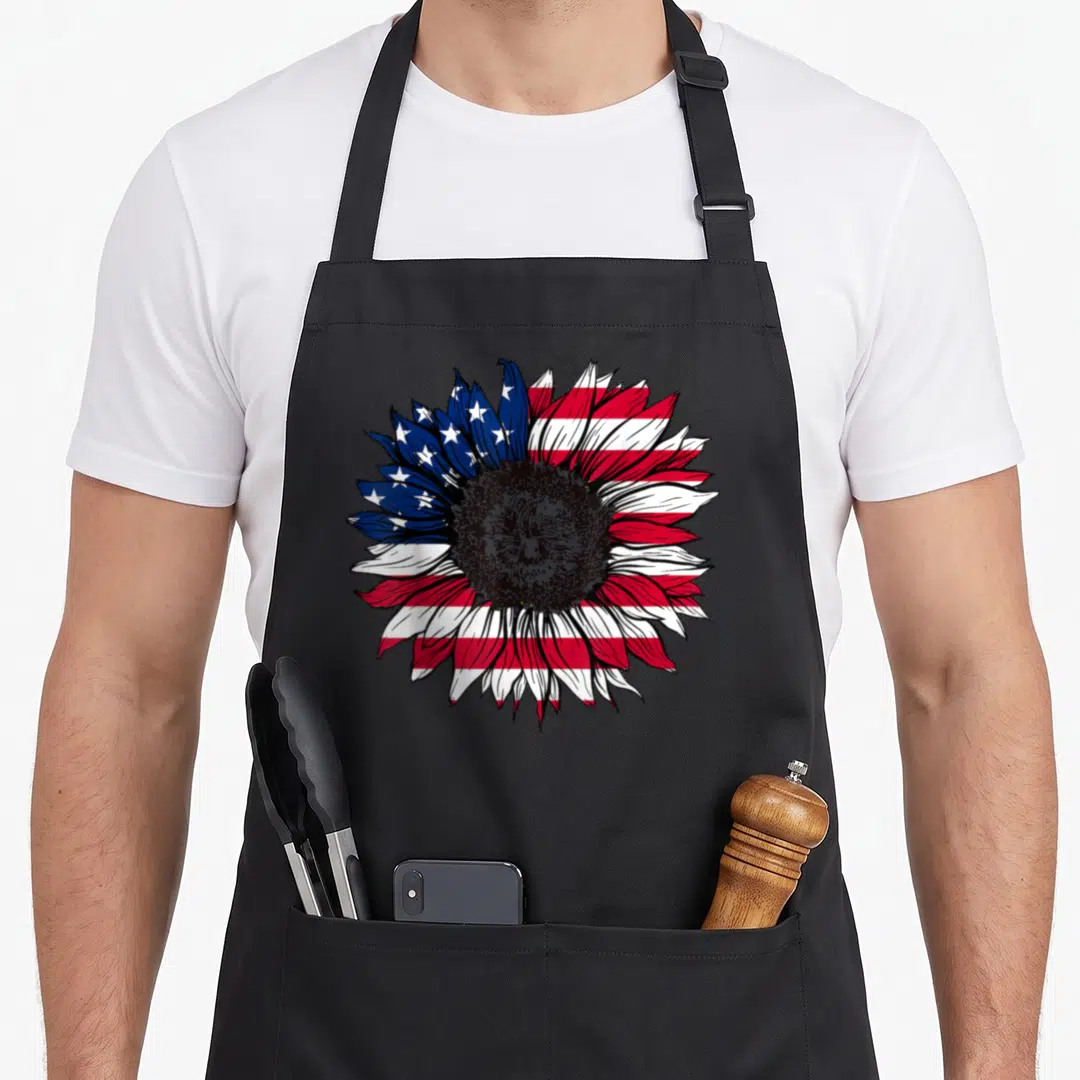 Flag sunflower 4th of july apron - Realistic sunflower with petals patterned in US flag blue white and red stripes — shown on a black cotton canvas apron against a white background