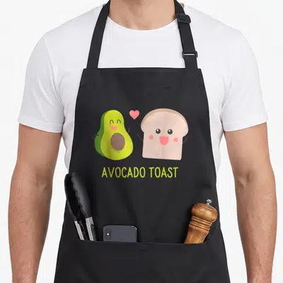 Cartoon toast apron - Cartoon avocado and toast with pink heart and AVOCADO TOAST text in green and brown — shown on a black cotton canvas apron against a white background
