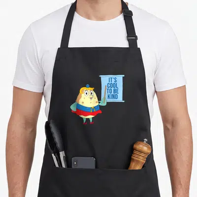 Kindness mrs apron - Cartoon pufferfish character holding blue banner with text IT'S COOL TO BE KIND — shown on a black cotton canvas apron against a white background