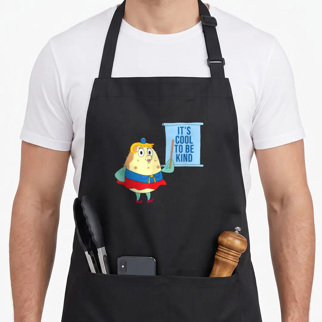 Kindness mrs apron - Cartoon pufferfish character holding blue banner with text IT'S COOL TO BE KIND — shown on a black cotton canvas apron against a white background