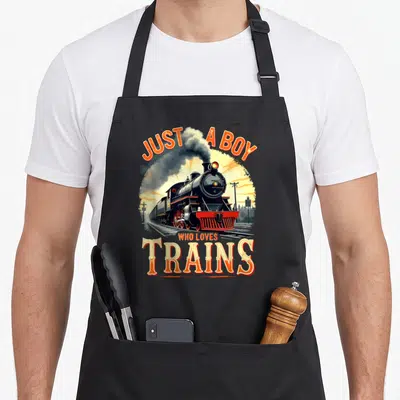 Vintage train steam locomotive apron - Retro steam locomotive illustration with orange JUST A BOY WHO LOVES TRAINS text — shown on a black cotton canvas apron against a white background