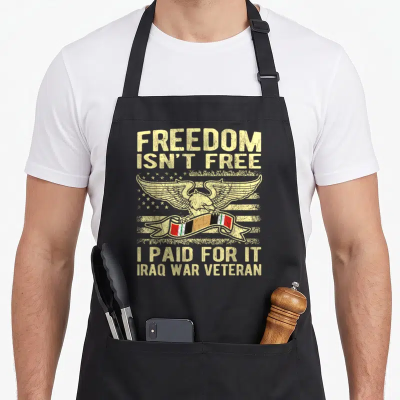 Freedom female veteran apron - Eagle over stars and stripes holding Iraqi flag ribbon with stacked beige text — shown on a black cotton canvas apron against a white background